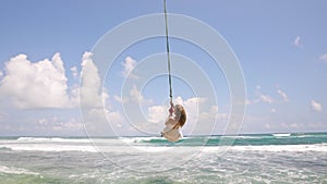 Woman in white dress enjoys swing ride above tropical beach waves. Blissful adult swings on rope by palm-fringed ocean