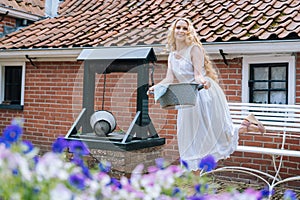 Woman in white dress holding basin near ancient artesian draw-well in village. Colorful violet flowers on foreground