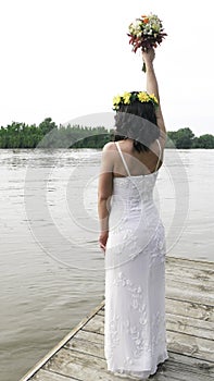 A woman in a white dress with a raised bouquet and flowers on her head looks at the river. Midsummer - Earth Day or Ivana Kupala.