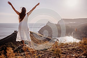 A woman in a white dress stands on a rocky hill overlooking the ocean. She is smiling and she is happy.