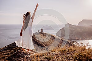 A woman in a white dress stands on a rocky hill overlooking the ocean. She is smiling and she is happy.