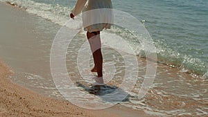 A woman in white dress walks barefoot on beach shoreline