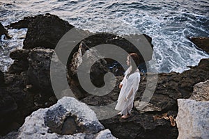 A woman in a white dress with wet hair stands on the stones by the ocean top view