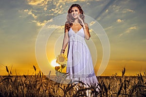 Woman in white dress in a wheat field with a sunflower flower at sunset