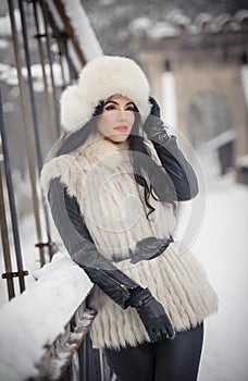 Woman with white fur cap and waistcoat enjoying the winter scenery near an iron fence. Attractive long hair brunette girl posing