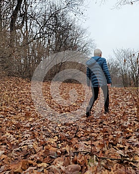 Woman with white hair and blue puffy jacket walks on fallen autumn leaves
