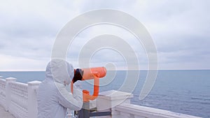 Woman in white jacket and hood looks in binoculars at sea waterfront with sea view. Side view.