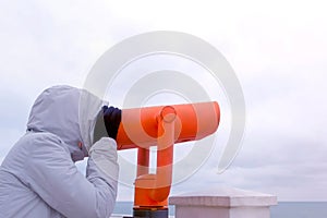 Woman in white jacket and hood looks in binoculars at sea waterfront with sea view. Side view.