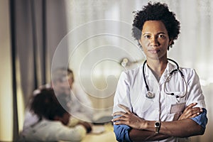 Woman in white lab coat with stethoscope standing in medical office for exam room at clinic looking at camera