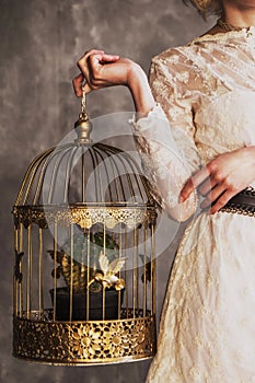 Woman in a white lace dress holding a golden cage with gold birds and a cactus. no face psychology therapy concept