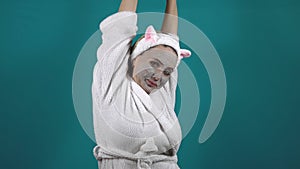 A woman in a white robe, wearing a carbonated bubble clay mask, yawns and stretches