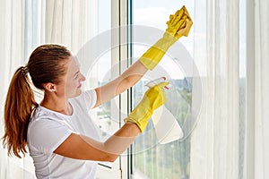 Woman in white shirt and yellow rubber gloves cleaning window with cleanser spray and yellow rag at home or office, copy space.