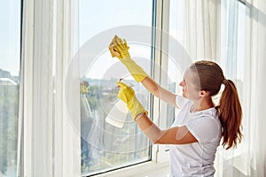 Woman in white shirt and yellow rubber gloves cleaning window with cleanser sprayer and yellow rag at home or office.