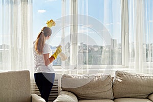 Woman in white shirt and yellow rubber gloves cleaning window with cleanser sprayer and yellow rag at home or office.