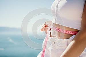 woman in white shorts holds in her hands pink measuring tape on her waist near the azure sea. Concept of detox and diet