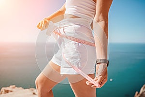woman in white shorts holds in her hands pink measuring tape on her waist near the azure sea. Concept of detox and diet