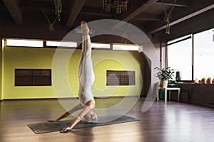 Woman in white sports jumpsuit practicing yoga performs shirshasana exercise, inverted asana pose, headstand