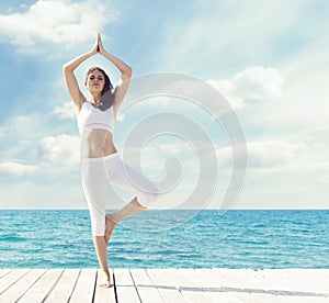 Woman in white sportswear doing yoga on a wooden pier. Sea and