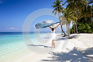 A woman in white summer dress walks on a tropical paradise beach with palm trees