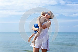 A woman in a white sundress with her little son in blue shorts laughs and hugs while walking by the sea on a sunny day. Love and