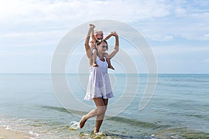 A woman in a white sundress with her little son in blue shorts laughs and hugs while walking by the sea on a sunny day. Love and