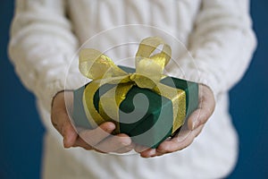 A woman in a white sweater is holding a green Christmas present on a blue background. Gift concept for Christmas and New Year