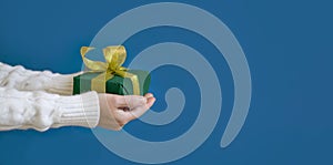 A woman in a white sweater is holding a green Christmas present on a blue background. Gift concept for Christmas and New Year