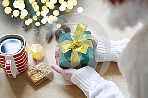 A woman in a white sweater holds a Christmas present on a wooden table background. Atmospheric photo with bokeh lights. Selective