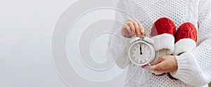 A woman in white sweater holds clock symbol of the new year and warm knitted red slippers. Concept of home cozy holiday