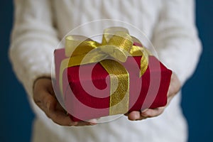 A woman in a white sweater holds a red Christmas present on a blue background. Gift concept for Christmas and New Year