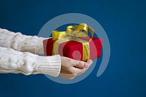 A woman in a white sweater holds a red Christmas present on a blue background. Gift concept for Christmas and New Year