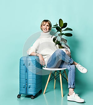 Woman in white sweater, jeans, sneakers. Smiling, holding ficus, sitting on chair, leaning on suitcase, posing on blue background