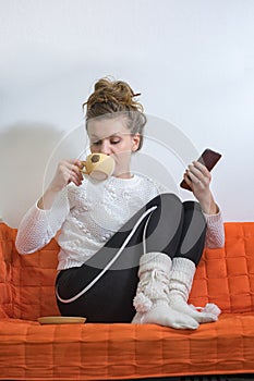 Woman in a white sweater and socks with pompons sits with legs on the sofa and drinks coffee, holding a smartphone photo