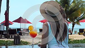 Woman in white swimwear and hat relax in swimming pool enjoy beautiful sea view with sun beds at sandy beach with