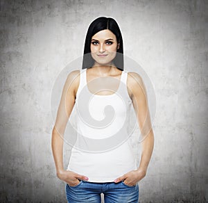 A woman in a white tank top and denims. Hands are in the pockets. Concrete background.