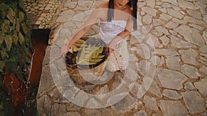 Woman in white top sitting on stone path holding a basket of bananas