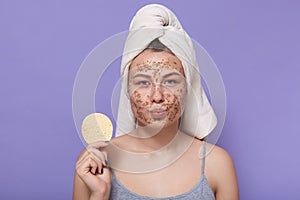 Woman with white towel on head, holding round cosmetic face sponge, being ready to removing facial mask off, peeling her face skin