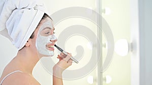 A woman in a white towel on her head applies a mask of blue clay to her face with a brush. Caring for the skin at home