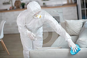 Woman in white workwear and protective gloves disinfecting furniture
