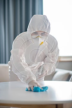 Woman in white workwear and protective gloves looking concentrated while disinfecting the table surface