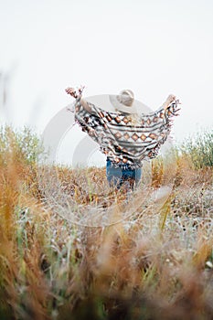 Woman in wide-brimmed felt hat and authentic poncho standing in high brown grass at foggy morning. Vertical orientation