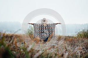 Woman in wide-brimmed felt hat and authentic poncho standing in high brown grass at foggy morning