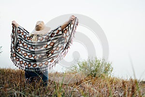 Woman in wide-brimmed felt hat and authentic poncho standing in high brown grass at foggy morning