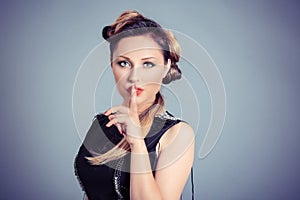 Woman wide eyed asking for silence or secrecy with finger on lips hush hand gesture blue grey background wall. Pretty girl placing