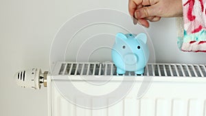 A woman in a winter bathrobe stands by the radiator and puts money into a piggy bank, Creative concept, High energy prices and hom