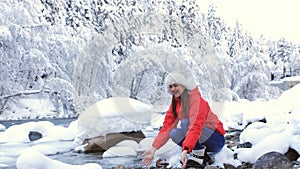 Woman in winter forest in the mountains sitting near a mountain stream and scoops hands icy water.