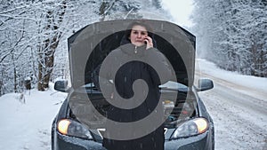 Woman in the winter forest is standing on the road with a broken car, opened the hood, talking on the phone, waiting for