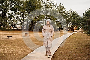 A woman in a winter jacket walks along a wooden path in a pine forest.