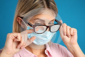Woman wiping foggy glasses caused by wearing disposable mask on blue background, closeup. Protective measure during coronavirus