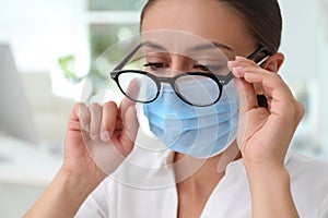 Woman wiping foggy glasses caused by wearing medical mask indoors, closeup